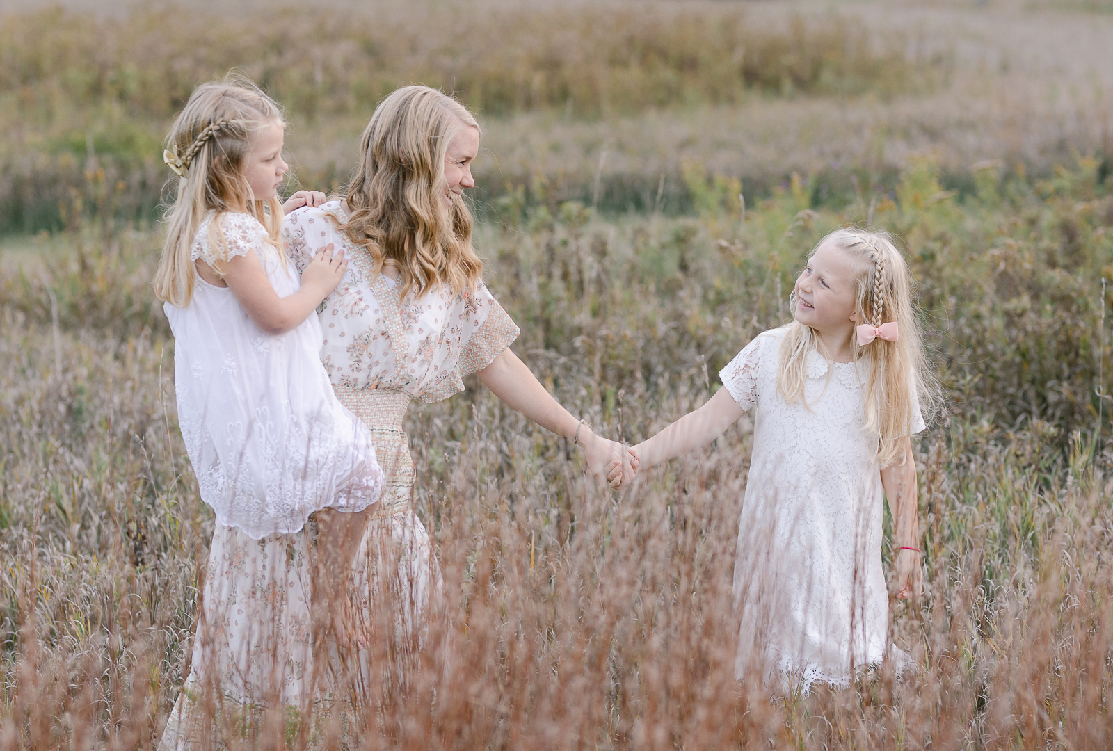 Detroit, Michigan family photographer, Laurel Smith Photography captures a mom holding her two daughter's hands while walking in tall grass. five year old girl white dresses, white Peter-pan collared little girl dress, tall grass family photos, bohemian ivory and white maxi dress for family pictures, little girl braids and bows, professional detroit michigan photographer #DetroitMichiganFamilyPhotographer #DetroitMichiganPhotographer #FamilyPhotoOutfitInspiration #PlymouthMichiganFamilyPhotographer #PlymouthMichiganPhotographer #ExtendedFamilyPhotos #DetroitMichiganFallFamilyPhotos #LetThemBeLittle #LetTheKids #DetroitMichiganLifestylePhotographer