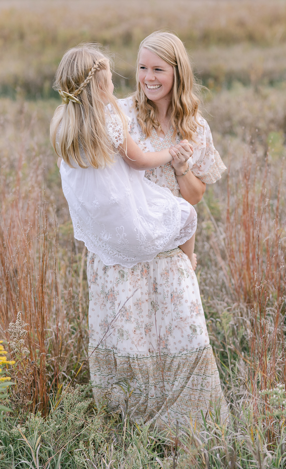 Plymouth, Michigan family photographer, Laurel Smith Photography captures a mom walking through a tall grass field while holding her daughter. tall grass family photos, plymouth michigan family photographer, ivory and white maxi dress with flutter sleeves, womens medium length blonde waved hair, little girl's half up braid with bow #DetroitMichiganFamilyPhotographer #DetroitMichiganPhotographer #FamilyPhotoOutfitInspiration #PlymouthMichiganFamilyPhotographer #PlymouthMichiganPhotographer #ExtendedFamilyPhotos #DetroitMichiganFallFamilyPhotos #LetThemBeLittle #LetTheKids #DetroitMichiganLifestylePhotographer