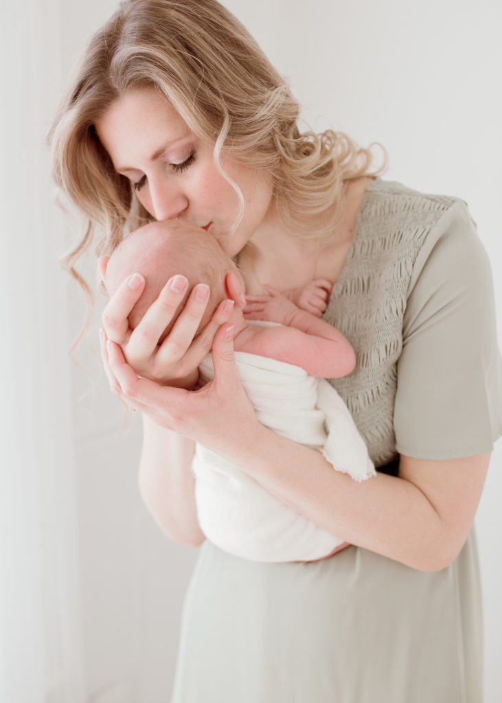 During this neutral colored, romantic-themed newborn photo session in Detroit, Michigan, Laurel Smith Photography captures this new mother holding hew newborn son and kissing him. mother kissing brand new baby boy, neutral colored newborn session, new mom, baby boy in loose white gauze swaddle, romantic newborn photos, detroit michigan newborn photographer #GreaterDetroitPhotographer #MichiganFamilyPhotographer #PlymouthMichiganPhotographer #PlymouthMichiganFamilyPhotographer #MichiganMaternityPhotos #StudioMaternityPhotos #Pregnant #ExpectantMother #NewbornPhotos #MichiganNewbornPhotographer