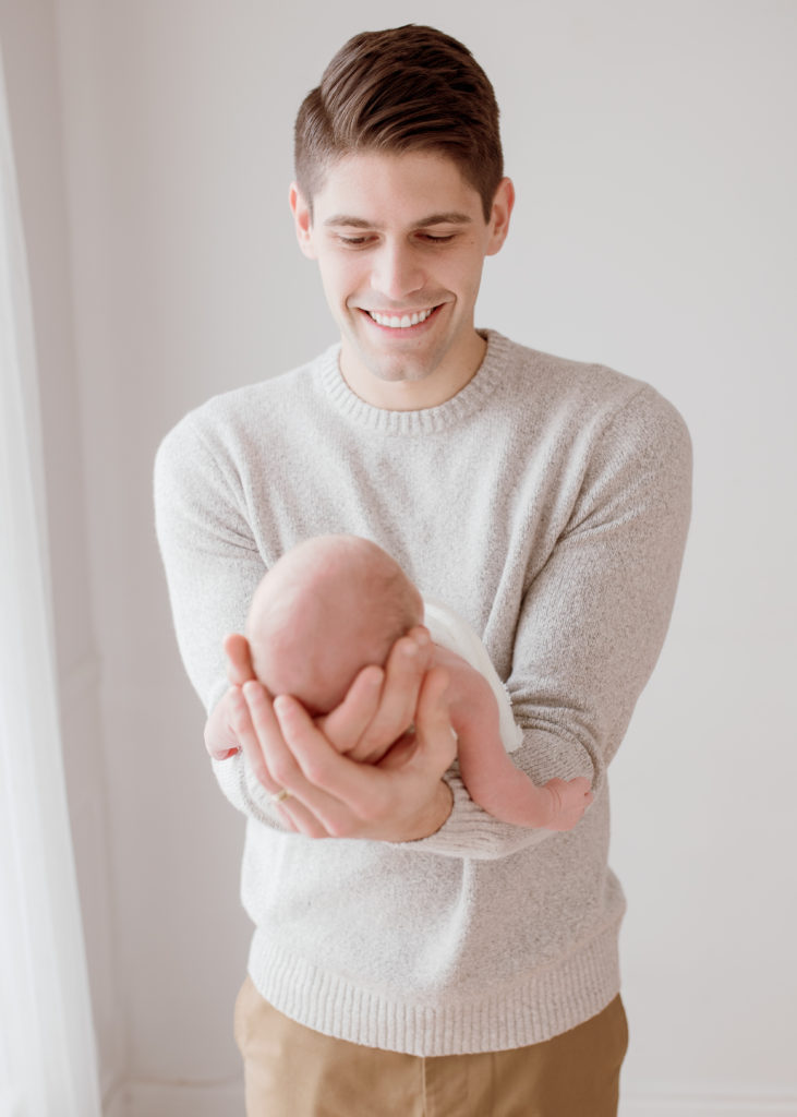 Posed in front of a natural light window at a photo studio in Plymouth, Michigan, Laurel Smith Photography captures this new father holding his newborn son in front of his tummy. newborn photographer plymouth michigan, laurel smith photography, neutral colored newborn photo session, cream mens sweater and khaki pants #GreaterDetroitPhotographer #MichiganFamilyPhotographer #PlymouthMichiganPhotographer #PlymouthMichiganFamilyPhotographer #MichiganMaternityPhotos #StudioMaternityPhotos #Pregnant #ExpectantMother #NewbornPhotos #MichiganNewbornPhotographer