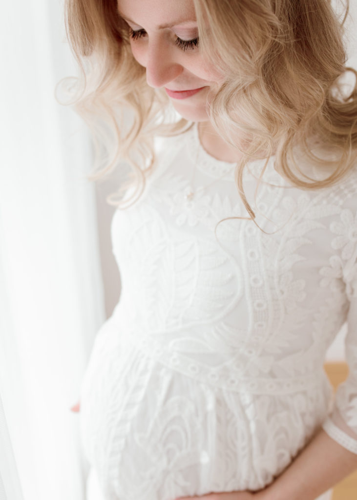 During this beautiful and neutral-colored maternity session in Greater Detroit, Michigan, Laurel Smith Photography captures up-close details. expectant mother looking down at baby bump, white lace embroidered maternity dress with elbow length sleeves, maternity photos posed in front of natural light windows with sheer white curtains, laurel smith photogrpahy, greater detroit maternity photographer #GreaterDetroitPhotographer #MichiganFamilyPhotographer #PlymouthMichiganPhotographer #PlymouthMichiganFamilyPhotographer #MichiganMaternityPhotos #StudioMaternityPhotos #Pregnant #ExpectantMother #NewbornPhotos #MichiganNewbornPhotographer