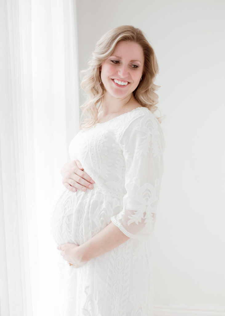 This expectant Plymouth, Michigan mother is posed in front of a natural light bearing window at this white photo studio during her maternity photoshoot with Laurel Smith Photography. womens medium length curled hair, womens white lace elbow-length maternity dress, natural light studio in plymouth michigan, plymouth michigan maternity photographer, pregnant #GreaterDetroitPhotographer #MichiganFamilyPhotographer #PlymouthMichiganPhotographer #PlymouthMichiganFamilyPhotographer #MichiganMaternityPhotos #StudioMaternityPhotos #Pregnant #ExpectantMother #NewbornPhotos #MichiganNewbornPhotographer