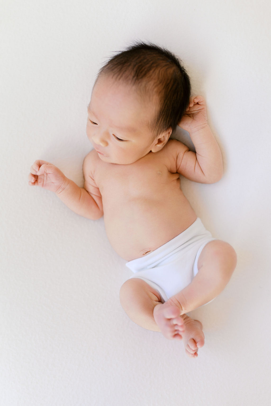 Newborn baby lying on a soft white backdrop wearing only a simple white wrap around the waist, one arm raised near the head and legs gently bent, with delicate skin tones and fine dark hair highlighted in soft natural light by the best newborn photographer bay area