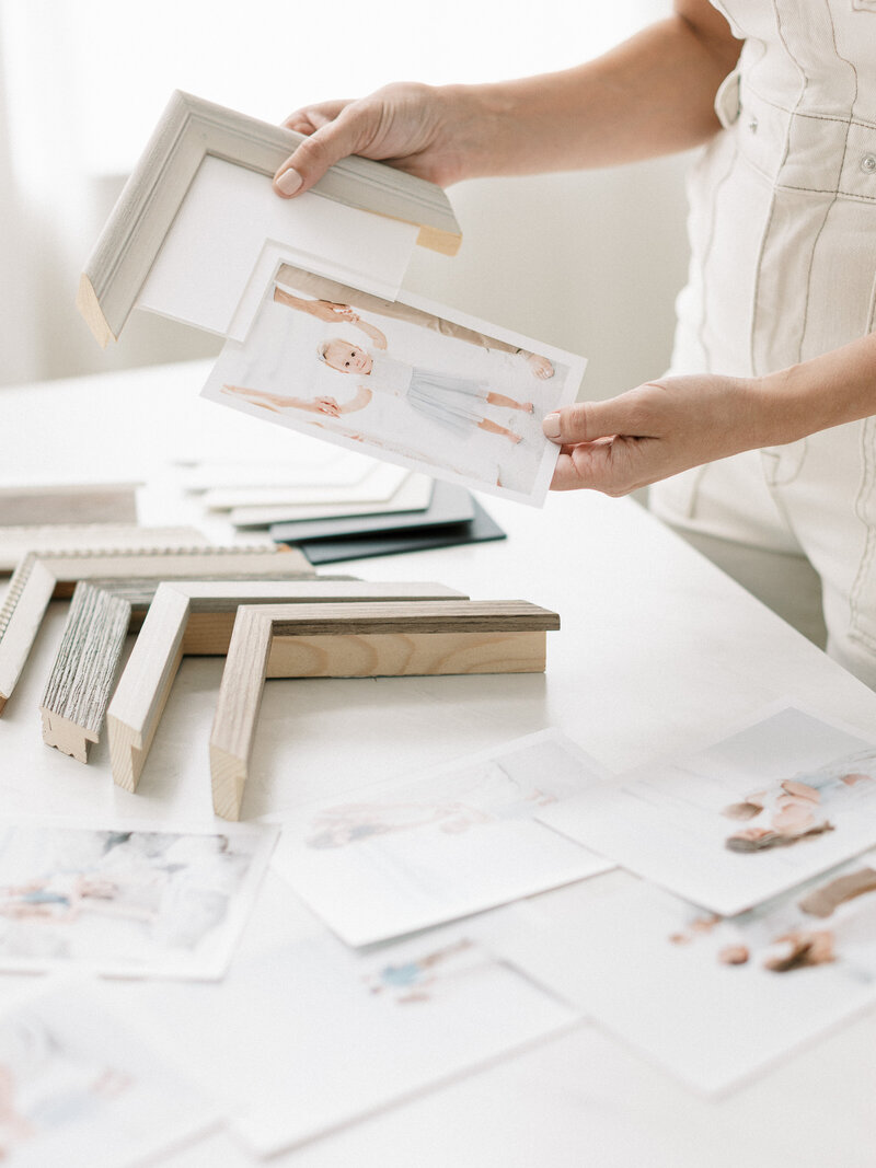 Hands arranging printed photographs and corner frame samples on a light-colored table, showing different frame finishes and small portrait prints laid out in a clean workspace, a helpful visual when learning how to choose a newborn photographer and display your images
