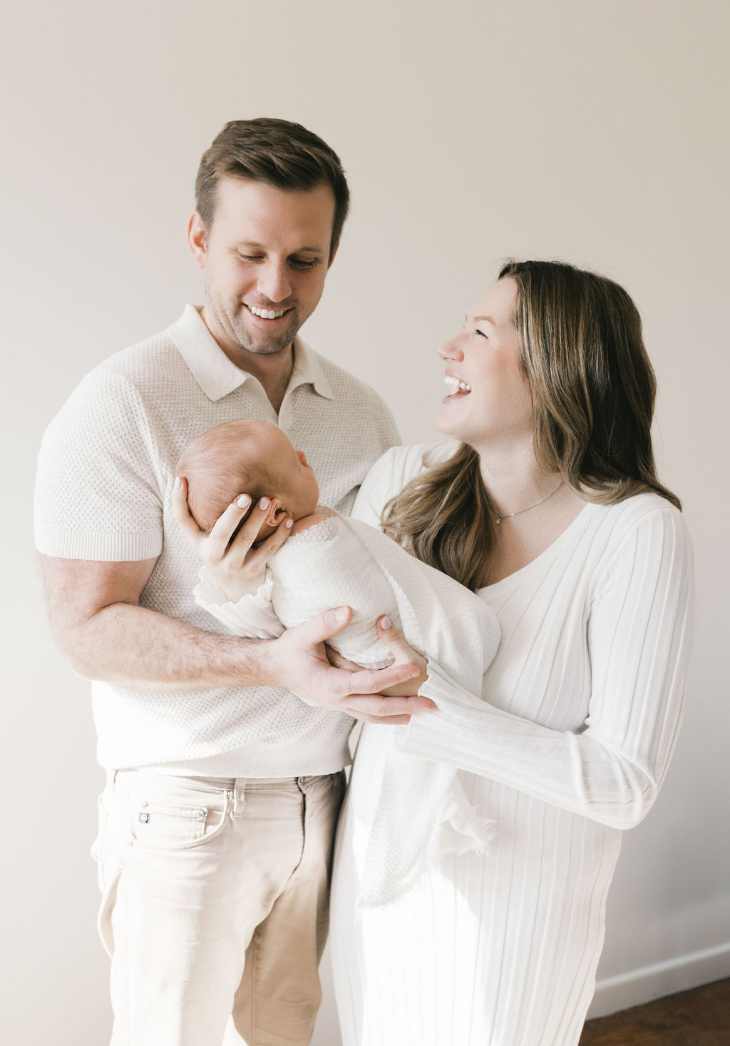 Smiling parents holding their newborn baby in a bright, minimal setting, sharing a joyful moment during a natural light lifestyle newborn photography session
