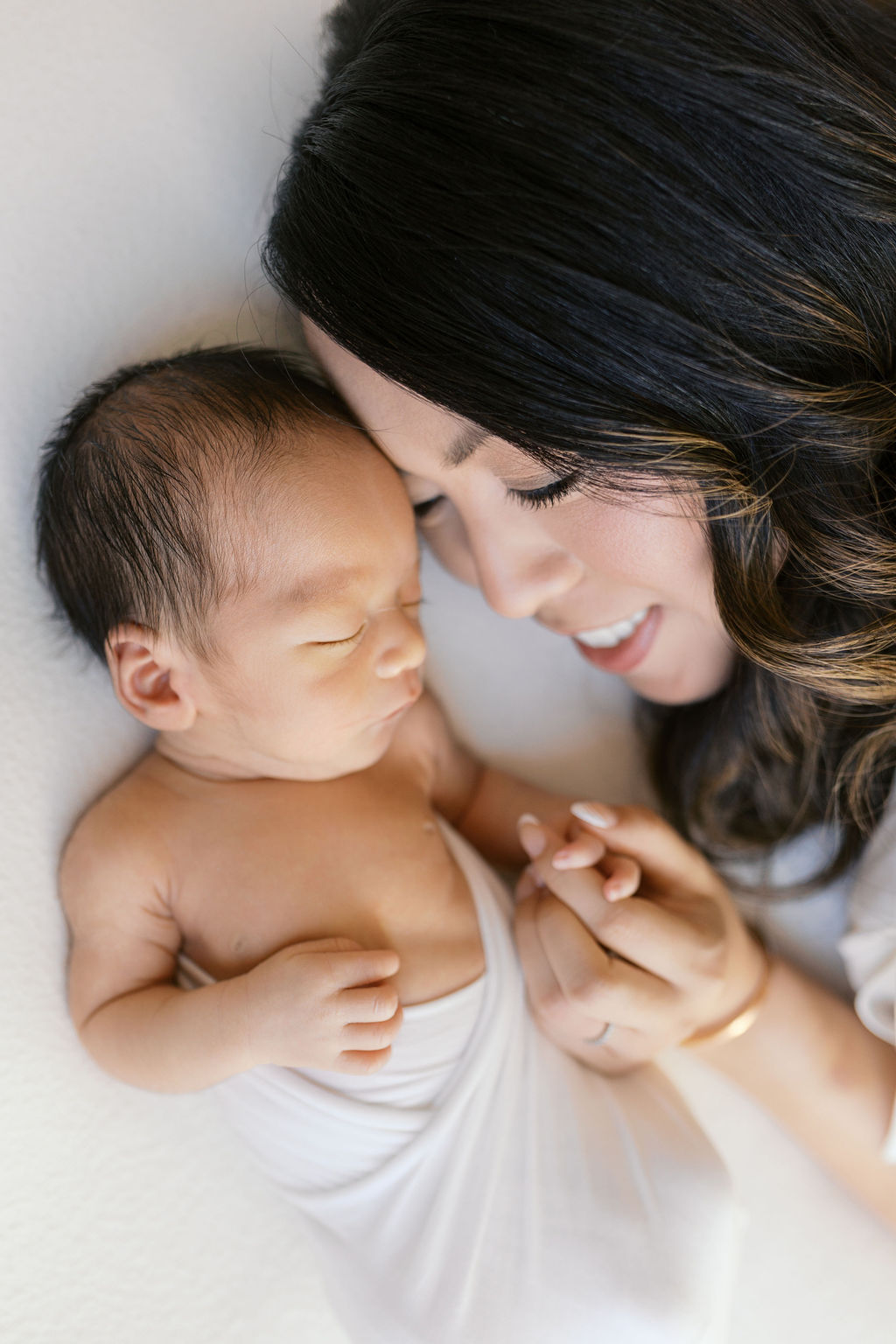 Mother leaning in close to her newborn, smiling softly as she holds the baby’s hand while the infant rests peacefully on a light background, with warm natural light emphasizing their bond in an intimate portrait by a bay area professional newborn photographer