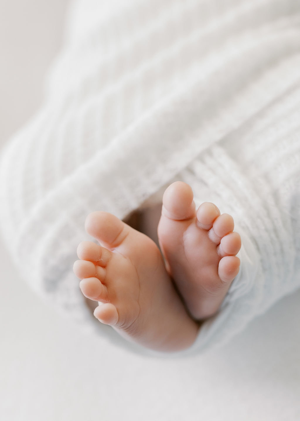 Newborn baby’s tiny feet peeking out from a soft white swaddle, toes slightly curled and softly lit to emphasize texture and warmth in a minimalist composition by a bay area professional newborn photographer