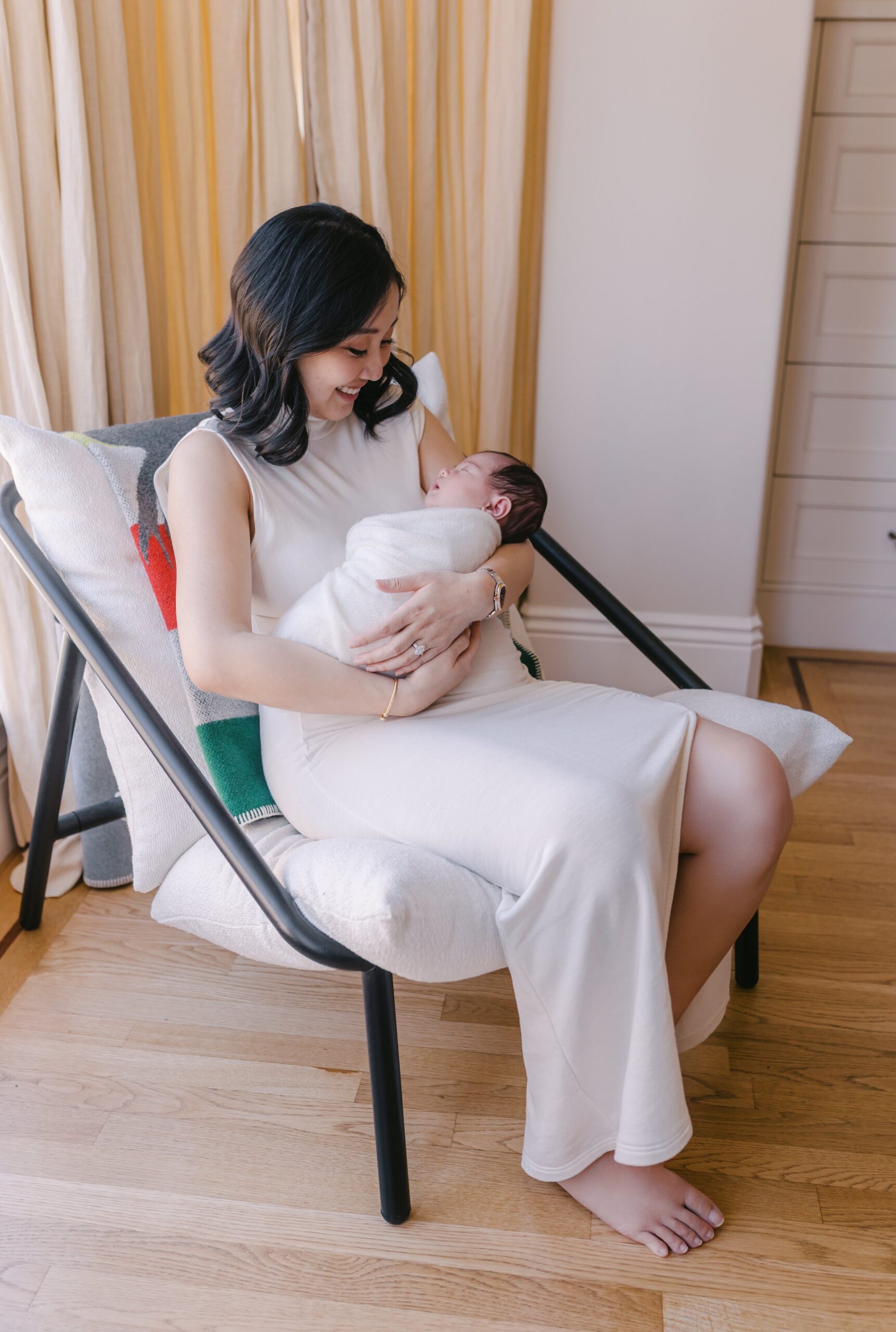 Mother seated in a modern cushioned chair with dark metal arms, smiling down at her swaddled newborn resting in her arms, soft beige curtains and warm wooden floors surrounding the scene in gentle natural light captured by a bay area newborn photographers session