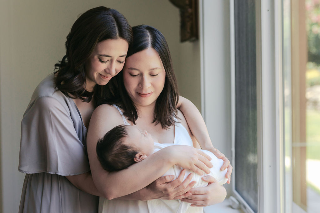 Two mothers standing close together beside a softly lit window, gently embracing as one cradles a newborn wrapped in white while both look down with calm, affectionate expressions, natural light illuminating their faces and the baby, a heartfelt family moment captured by the best newborn photographer bay area