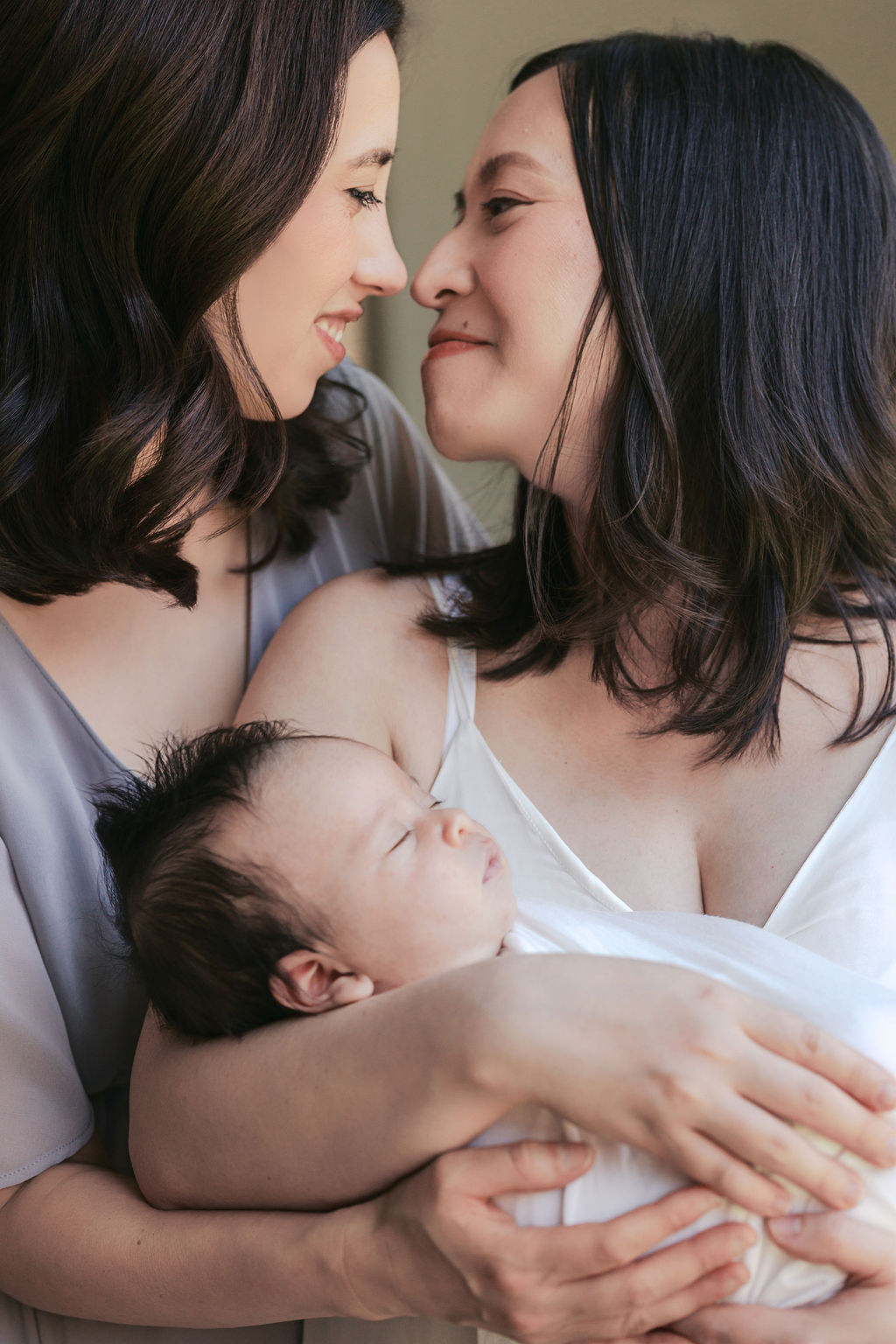Two mothers holding their newborn close between them, gazing at each other with soft smiles while the baby rests peacefully in their arms, warm natural light emphasizing connection in a moment captured by a bay area professional newborn photographer