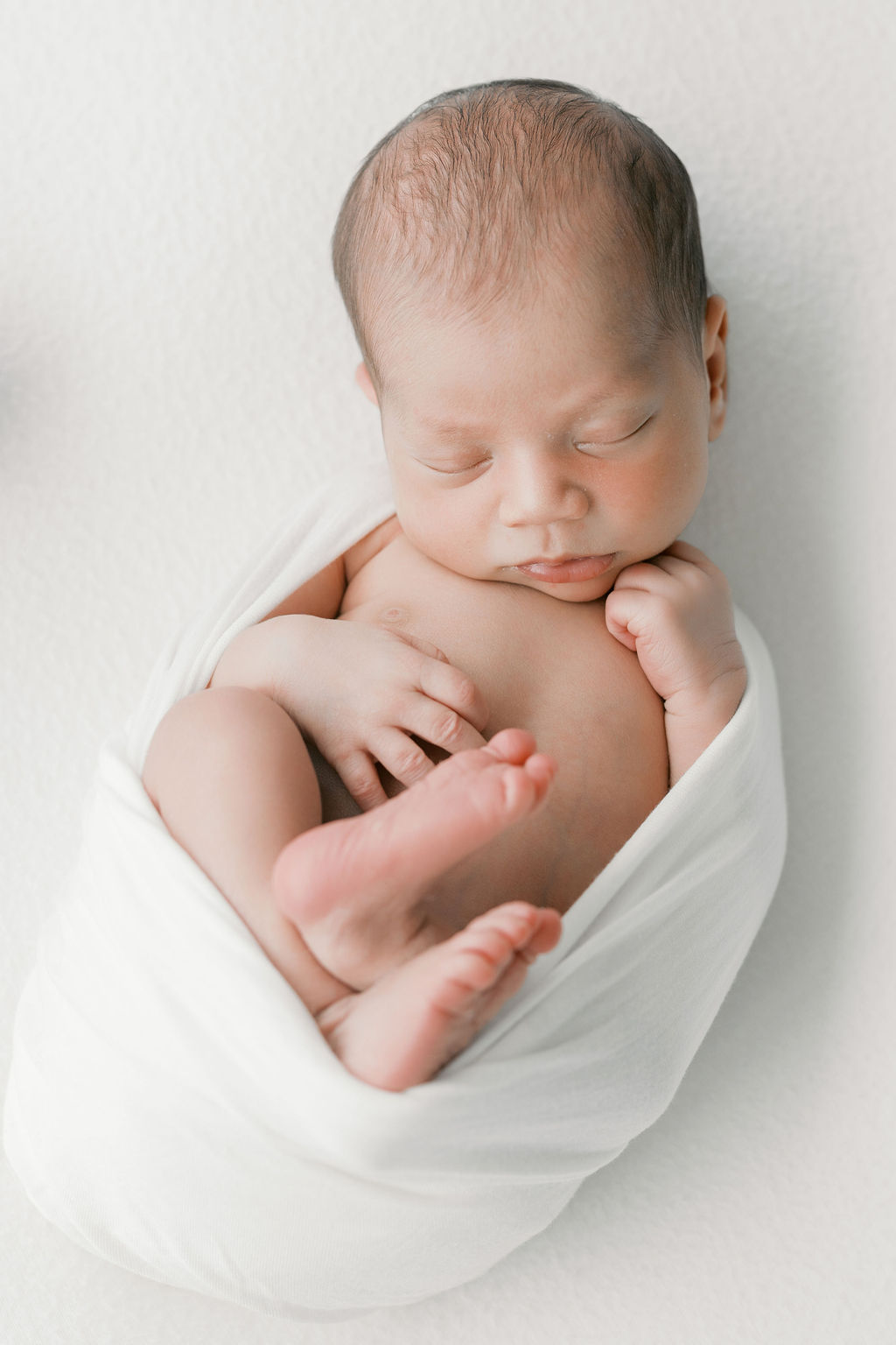 Newborn baby peacefully sleeping in a curled pose on a light neutral surface, loosely wrapped in a white swaddle with small feet and hands visible, soft shadows and creamy tones highlighting texture in an image by the best newborn photographer bay area