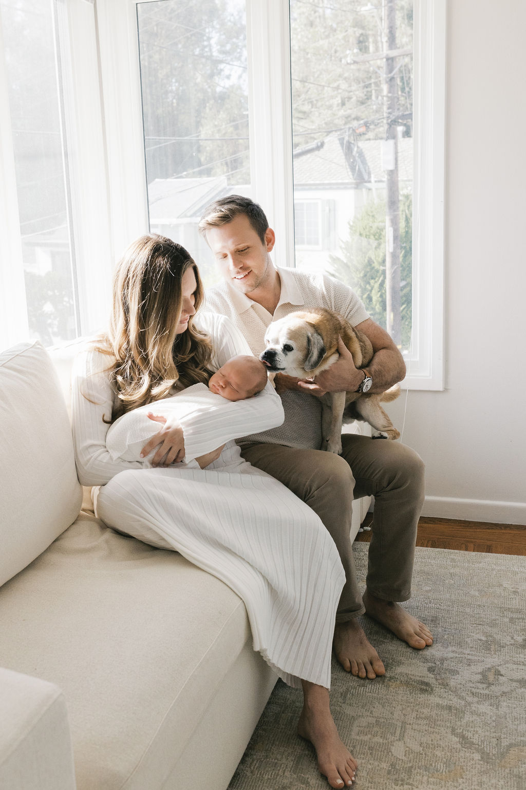 Mother and father sitting close together on a light-colored sofa in a sunlit room, with the mother gently cradling a sleeping newborn wrapped in a white swaddle while the father holds a small dog beside them, soft natural light streaming through large windows during a bay area newborn photographers lifestyle session