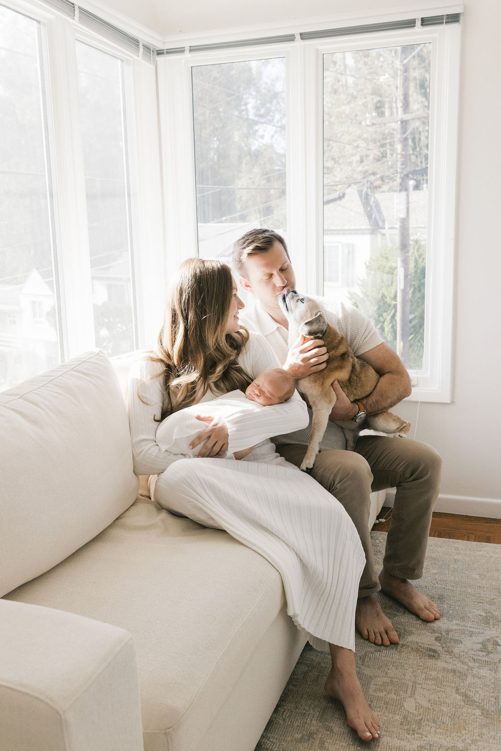 Parents seated on a neutral sofa near bright windows, smiling softly as the father leans in to kiss their small dog while the mother holds their peacefully sleeping newborn in a white wrap, capturing a warm family moment by a bay area professional newborn photographer