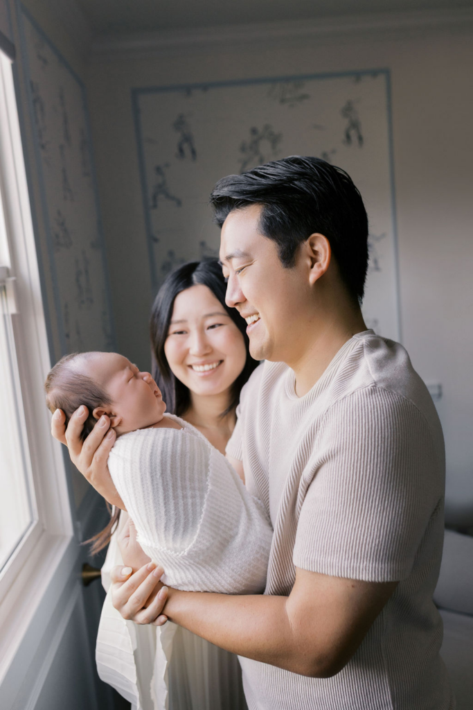Father standing beside a bright window holding a swaddled newborn up at chest height, smiling warmly while the mother stands just behind him watching, soft natural light highlighting the baby’s profile in a tender moment photographed by the best newborn photographer bay area