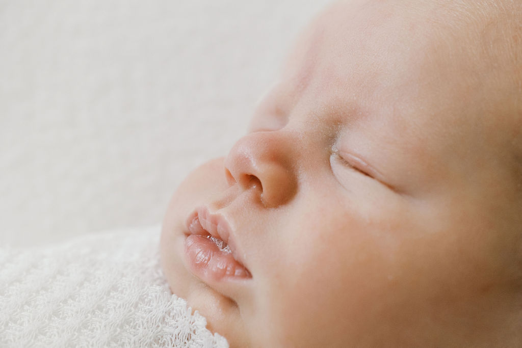 Close-up side profile of a sleeping newborn’s face resting against a soft white blanket, with delicate lips slightly parted and smooth skin gently illuminated by natural light, captured by the best newborn photographer bay area