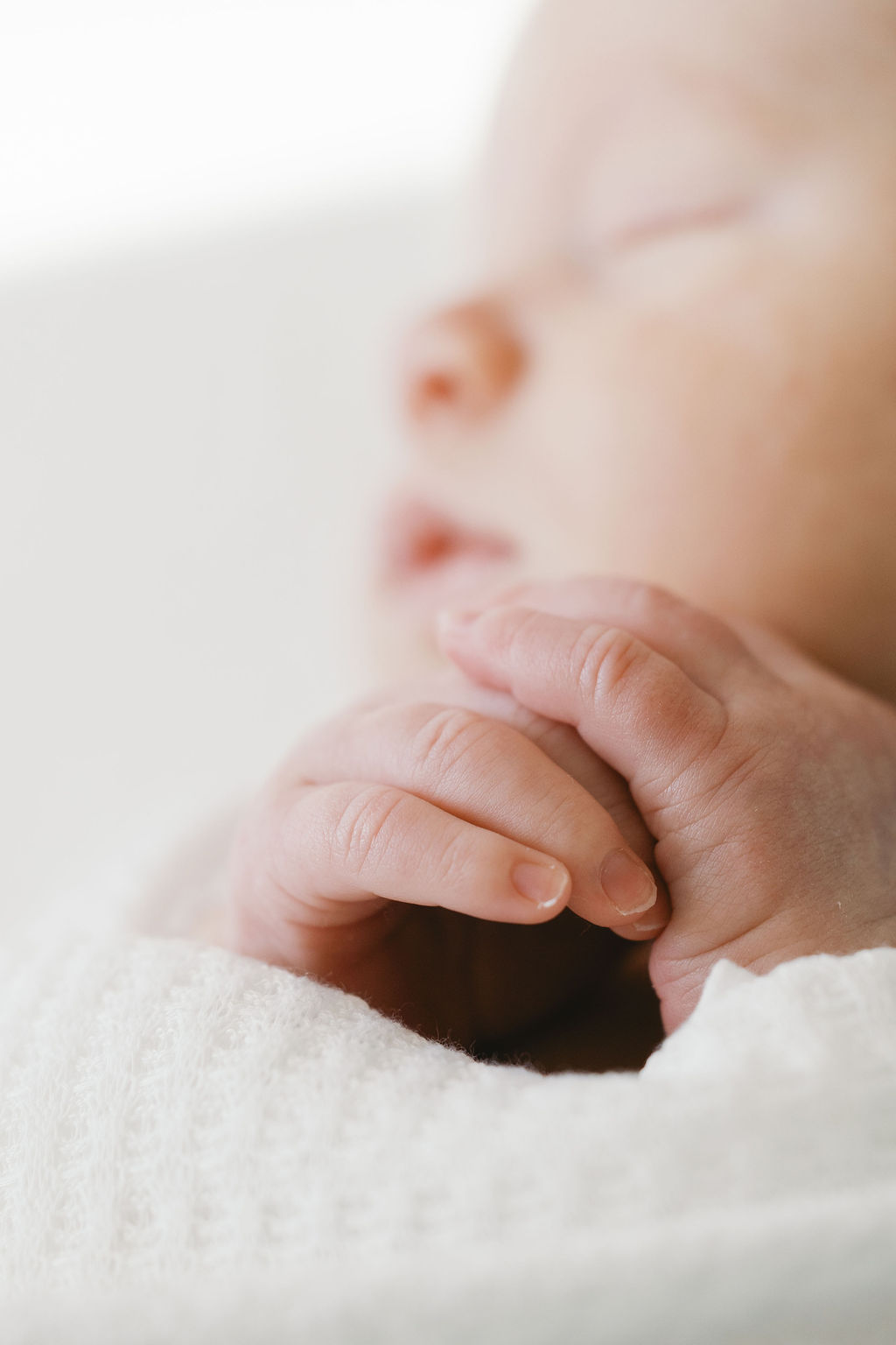 Macro detail of a newborn baby’s tiny hands softly clasped together near the face, wrapped in a white blanket with a shallow depth of field emphasizing texture and tenderness in a portrait by bay area newborn photographers
