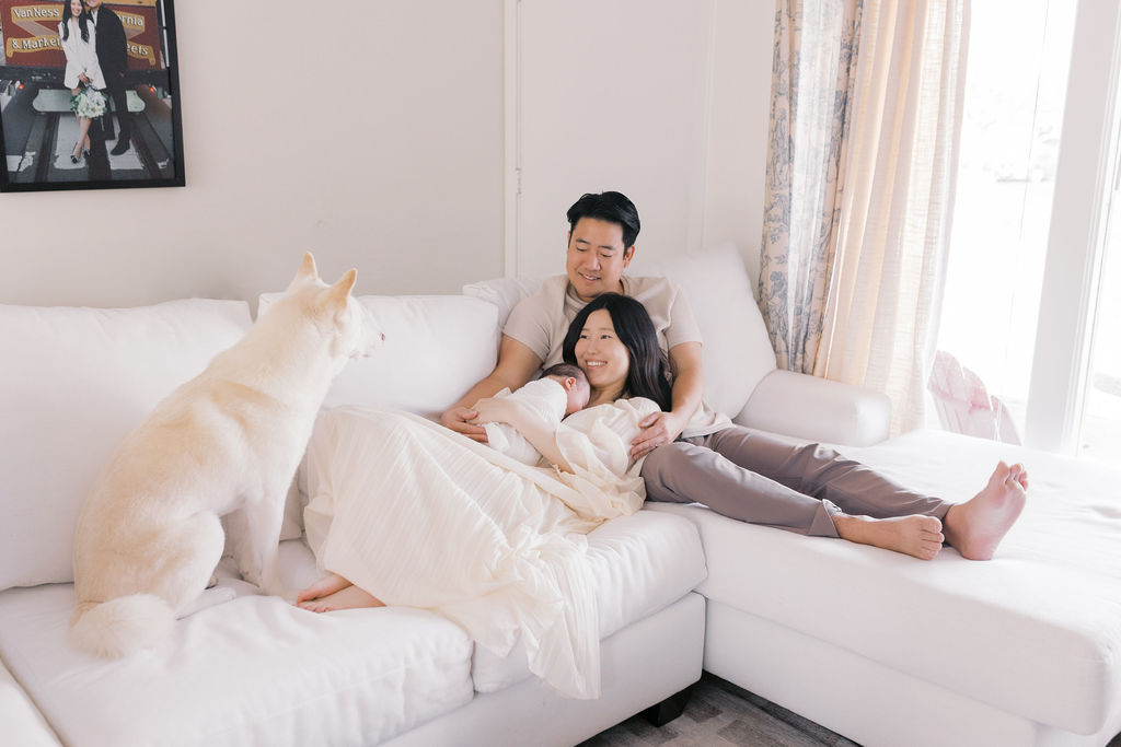 New parents relaxing on a bright white couch with their newborn sleeping in the mother’s arms while a large white dog sits nearby watching them, soft window light filling the room and highlighting a peaceful family moment captured by the best newborn photographer bay area