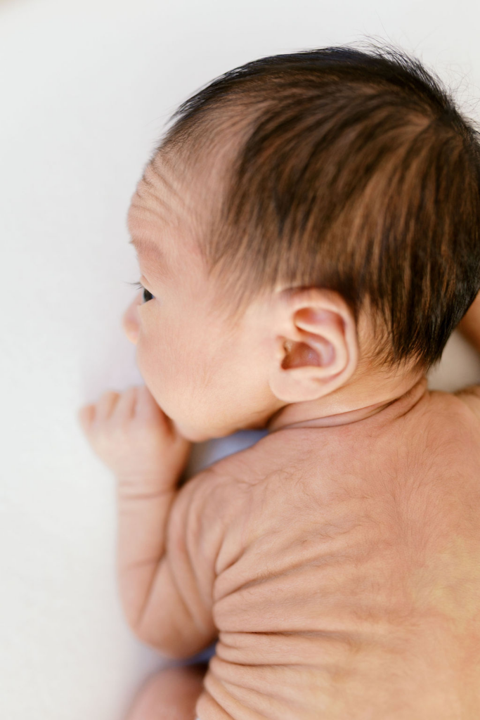 Side profile of a newborn baby lying on their stomach with fine dark hair, soft wrinkles along the back and shoulders, and a tiny hand tucked near the face, captured in gentle natural light during a best newborn photographer bay area session