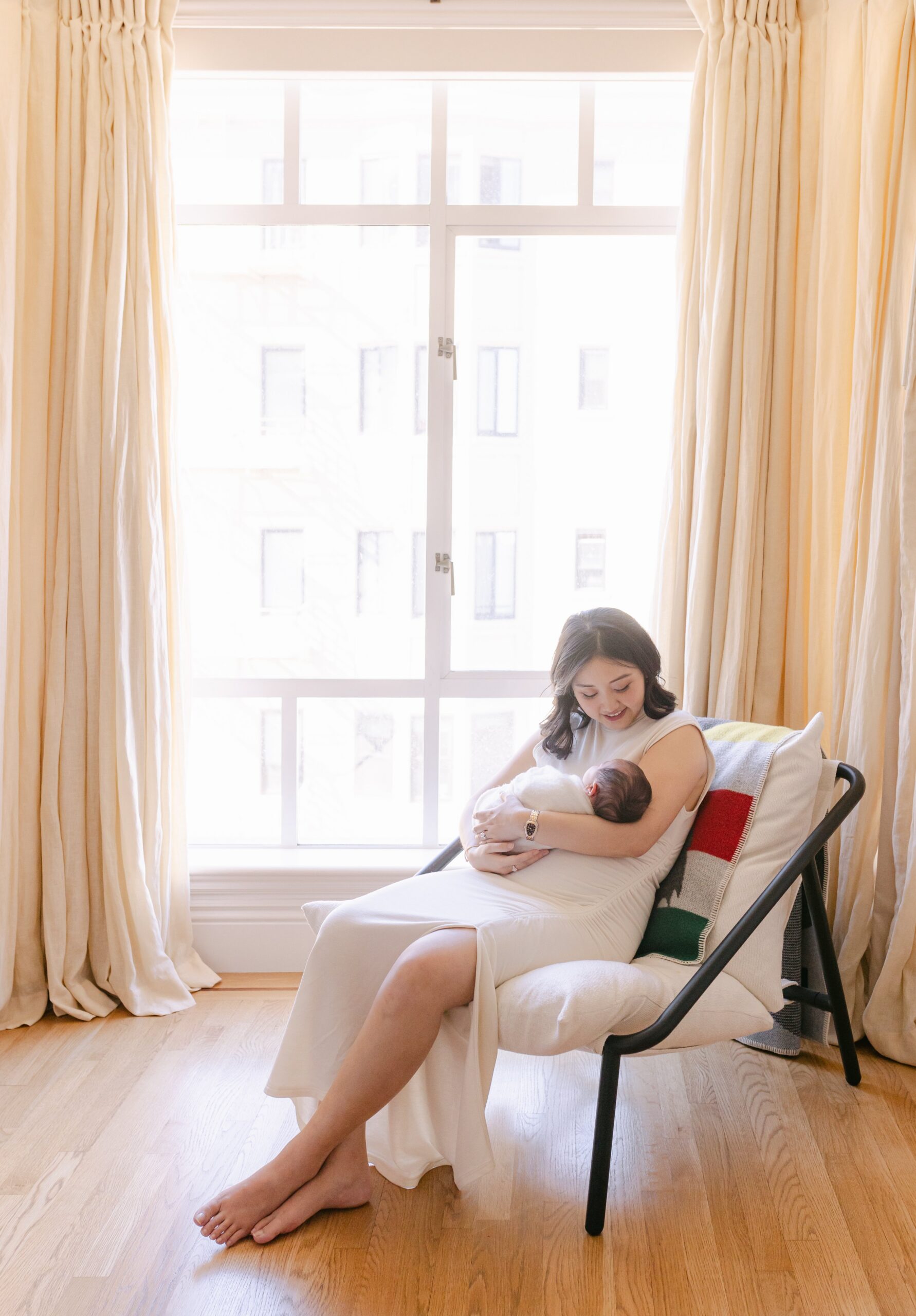 Wide view of a mother sitting in a chair near a large bright window framed by soft cream curtains, cradling her newborn wrapped in white while looking down with a calm smile, illustrating the soft natural style of a bay area professional newborn photographer