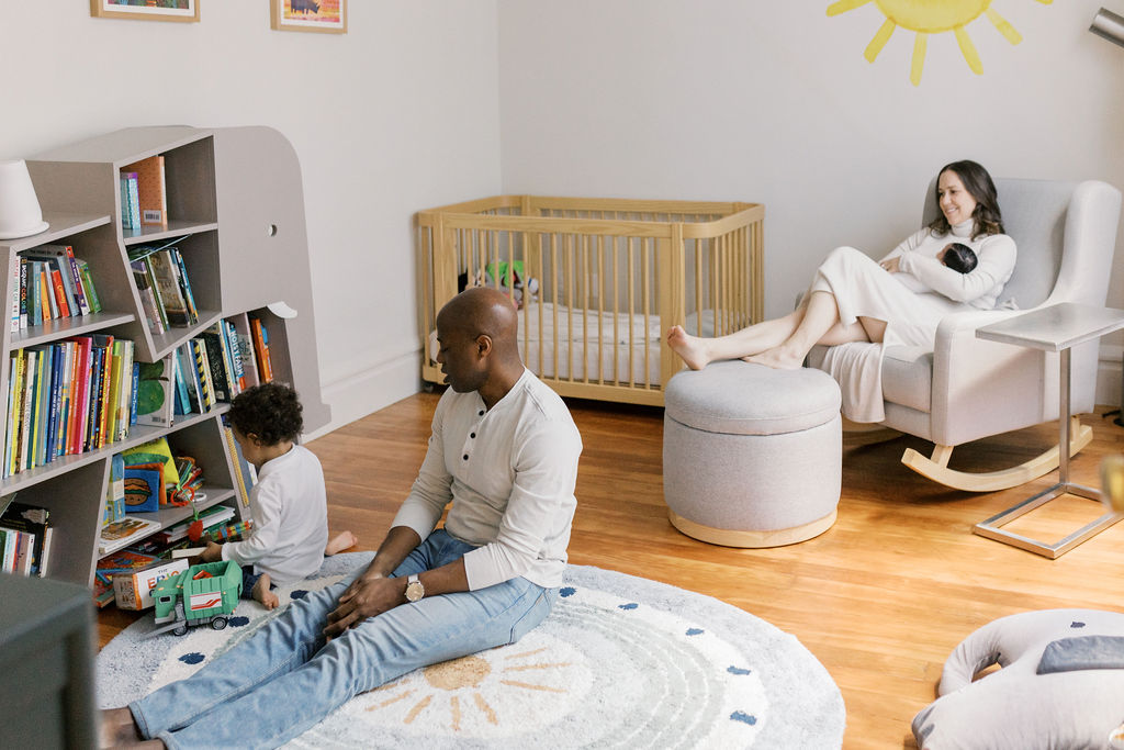 Wide view of a bright nursery with wooden floors, where a father sits on a soft patterned rug beside a toddler playing with a toy truck near a low bookshelf filled with children’s books, while in the background a mother relaxes in a chair cradling a sleeping newborn near a wooden crib, a natural in-home moment captured by bay area newborn photographers