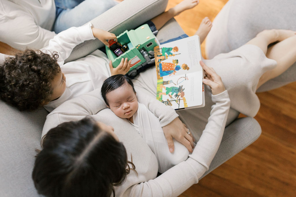 Overhead view of a mother sitting in a soft chair holding a peacefully sleeping newborn wrapped in white, while an older sibling with curly hair sits beside her playing with a toy truck and looking at a colorful picture book, a candid everyday moment captured by bay area newborn photographers