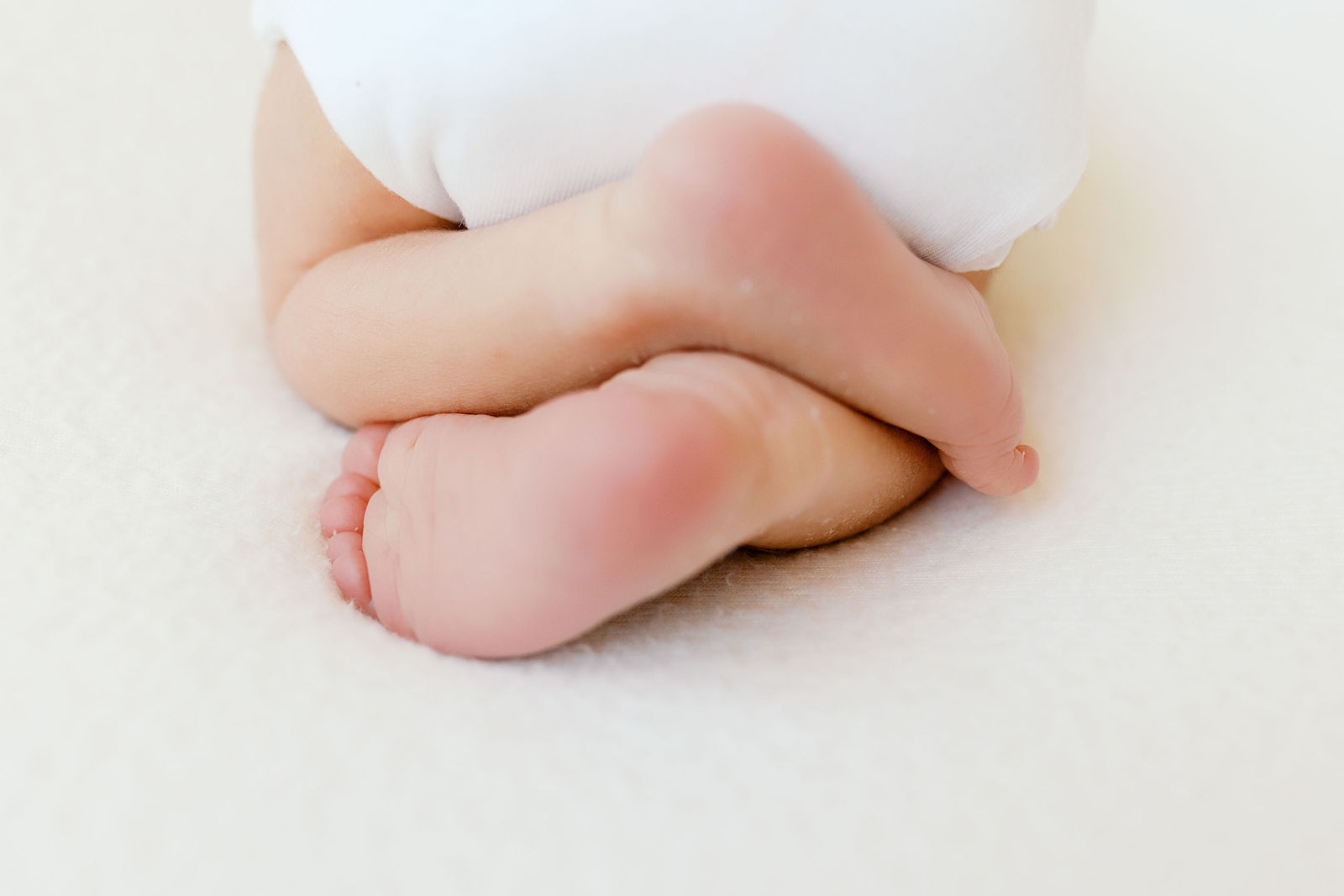 Close-up of a newborn baby’s feet gently crossed at the ankles, with soft pink soles and tiny curled toes resting on a plush white blanket, subtle skin texture visible in soft natural light, a detail often captured by the best newborn photographer bay area