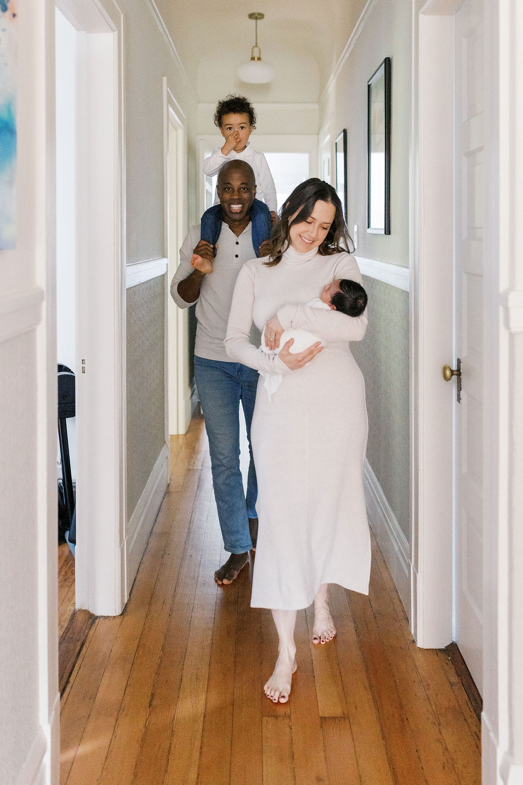 Family walking barefoot down a softly lit hallway with warm wooden floors, the mother cradling a swaddled newborn while the father follows behind carrying their toddler on his shoulders, a candid in-home moment that reflects how to choose a newborn photographer who captures natural connection and movement