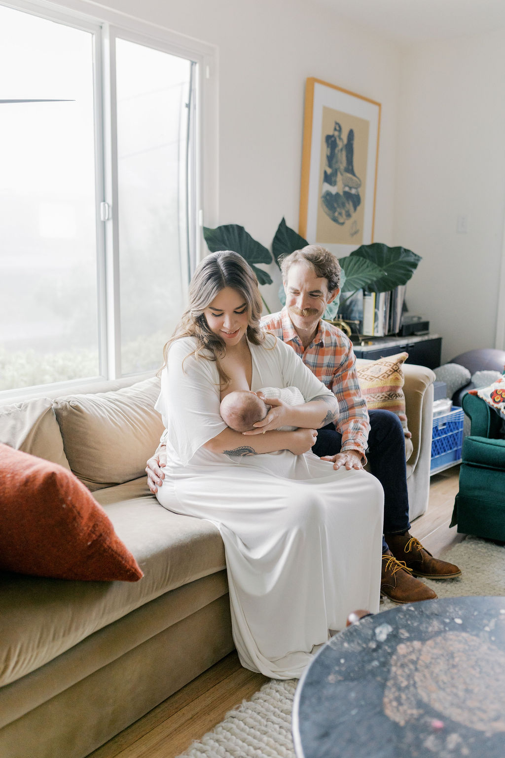 Mother seated on a neutral sofa gently cradling her newborn while the father sits close beside her with a hand resting supportively on her back, soft daylight coming through a nearby window and houseplants and artwork adding warmth to the space, a relaxed in-home moment captured by bay area newborn photographers