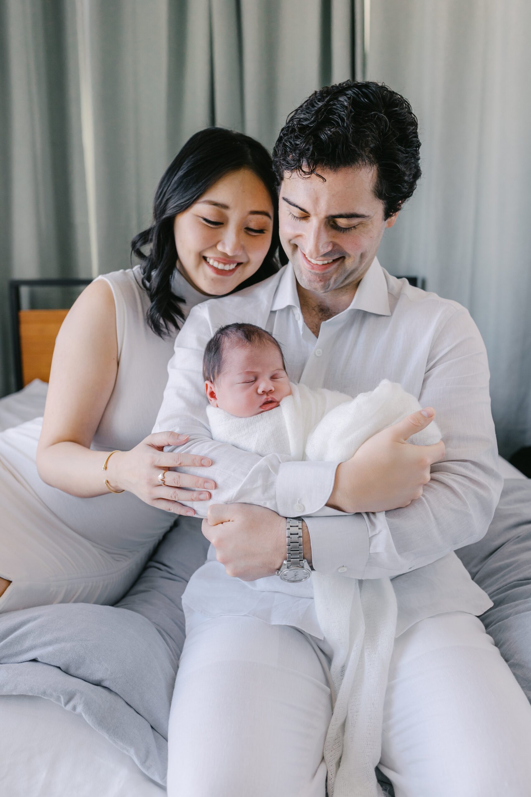 Smiling parents sitting on a bed, gently cradling their swaddled newborn baby while looking down with joy in a cozy, light-filled lifestyle newborn photography session in the Bay Area
