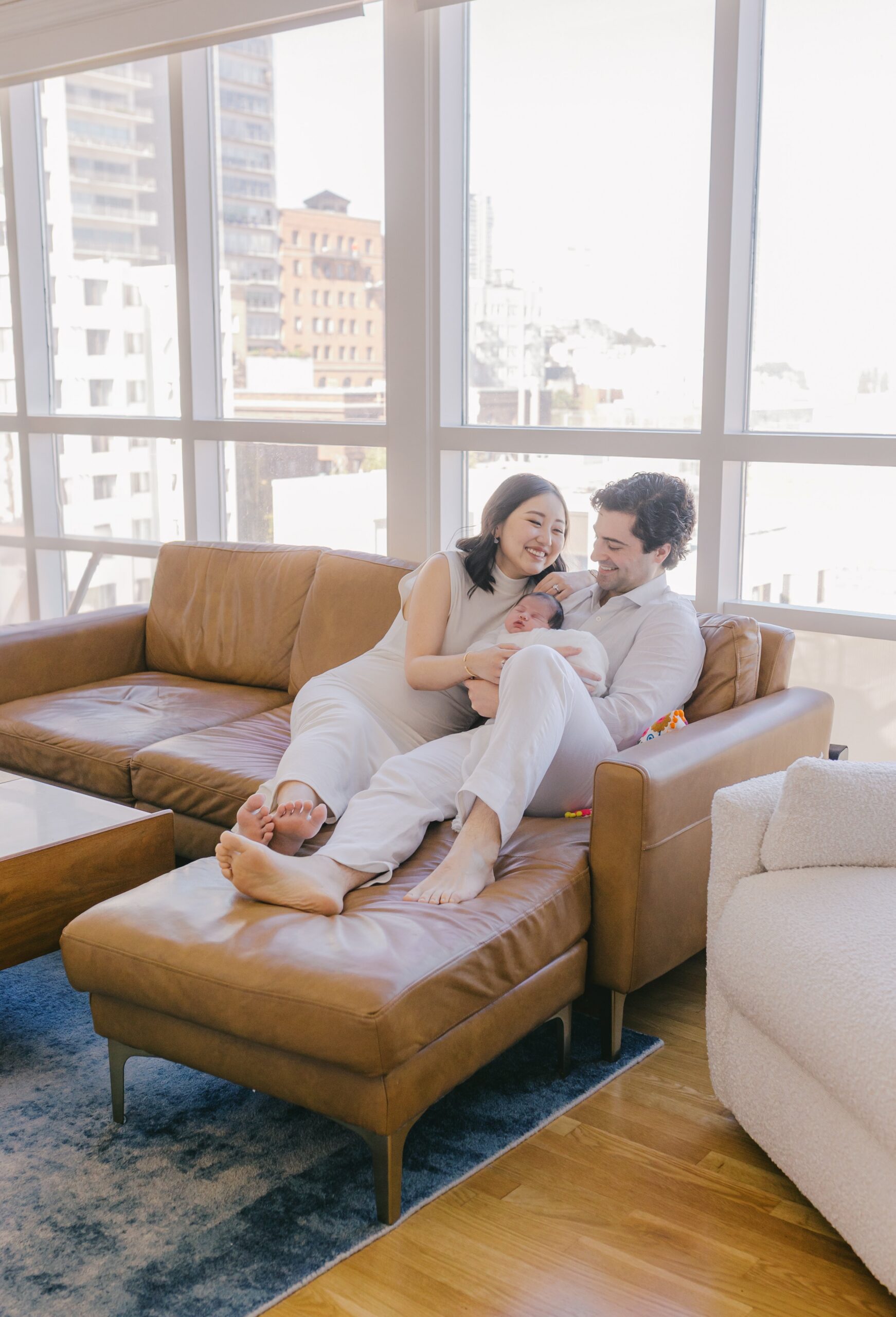 Parents sitting closely together on a tan leather sofa with large sunlit windows behind them, smiling as they hold their newborn nestled between them, city buildings softly visible outside, a relaxed family moment captured by the best newborn photographer bay area