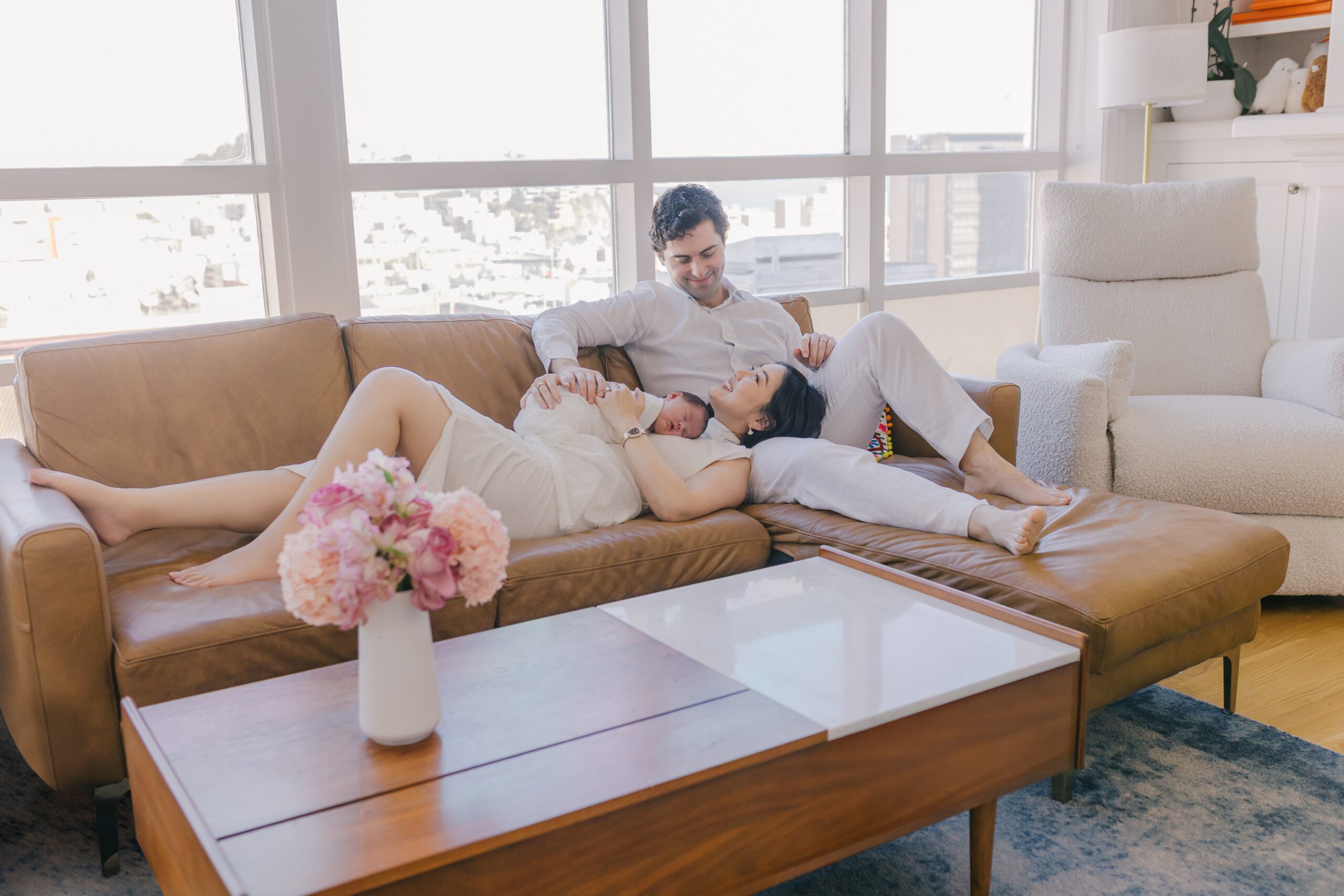 Parents relaxing together on a tan leather sofa in a bright living room with large windows, the mother lying back with a sleeping newborn resting on her chest while the father sits beside her smiling down, a vase of pink flowers on the coffee table in the foreground and soft city views beyond, captured by bay area newborn photographers