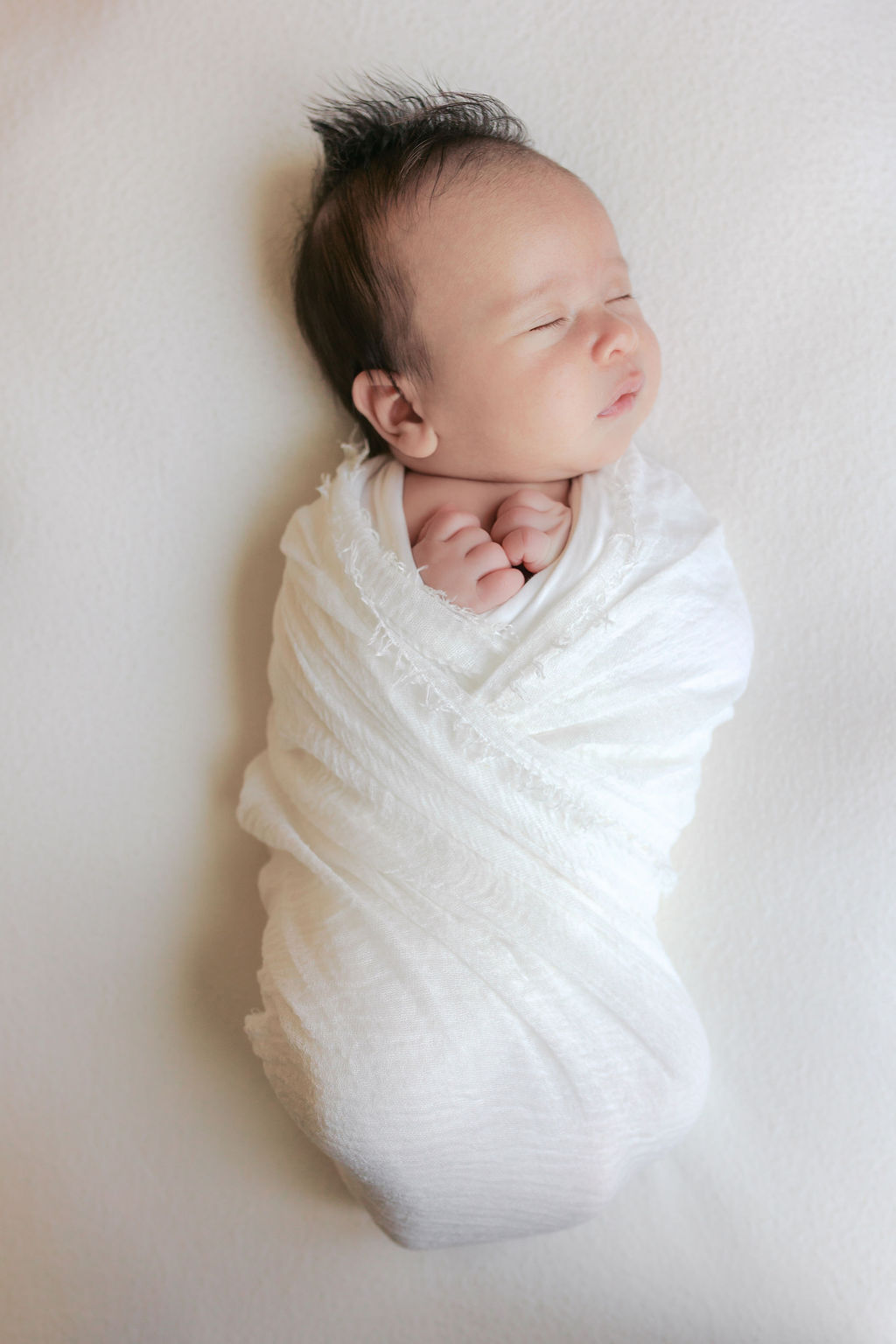 Newborn baby tightly wrapped in a soft white swaddle, lying on a neutral textured surface with tiny hands tucked under the chin and wisps of dark hair visible, softly lit to highlight delicate features by the best newborn photographer bay area