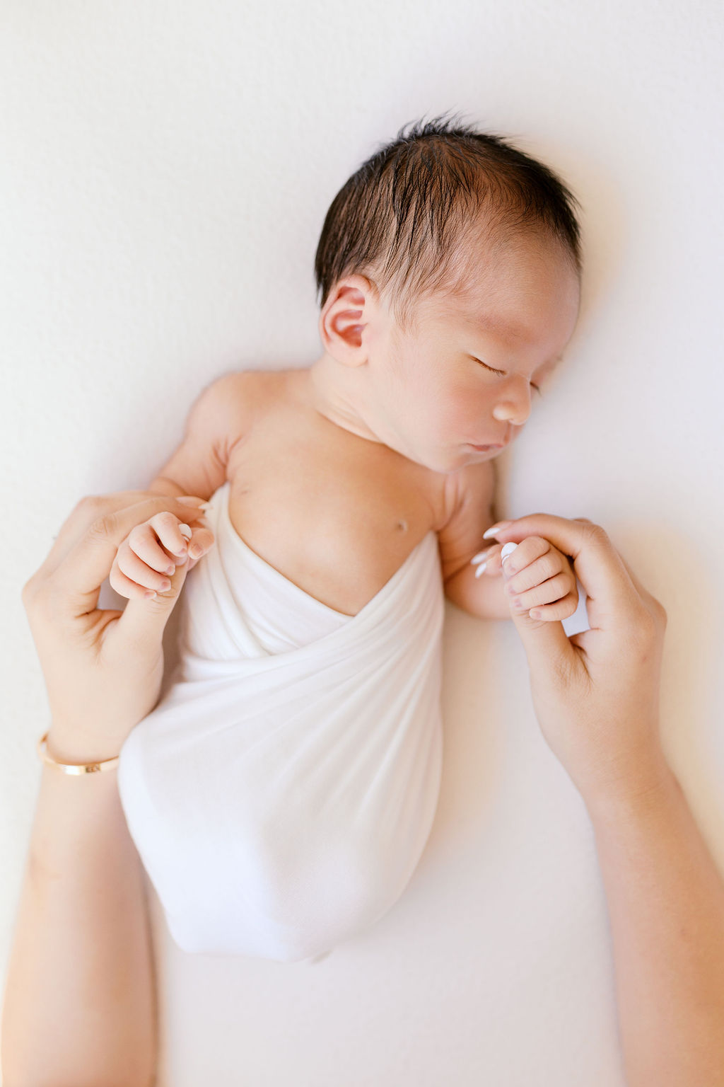 Sleeping newborn wrapped snugly in a white swaddle, lying on a neutral background while a parent gently holds both of the baby’s tiny hands, creating a sense of scale and connection in a calm, softly lit scene captured by bay area newborn photographers