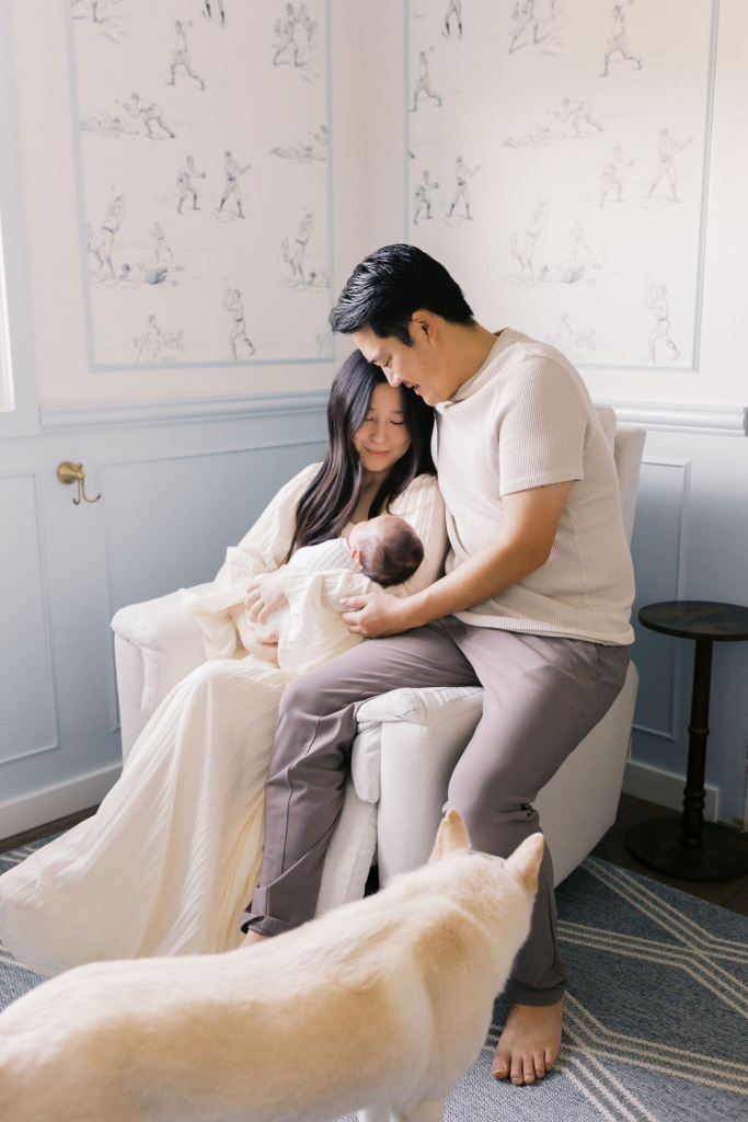 Mother and father seated closely together in a cushioned chair, gently leaning their heads together as they look down at their newborn resting in the mother’s arms, while a light-colored dog stands nearby in the foreground, soft window light illuminating the scene captured by a bay area professional newborn photographer