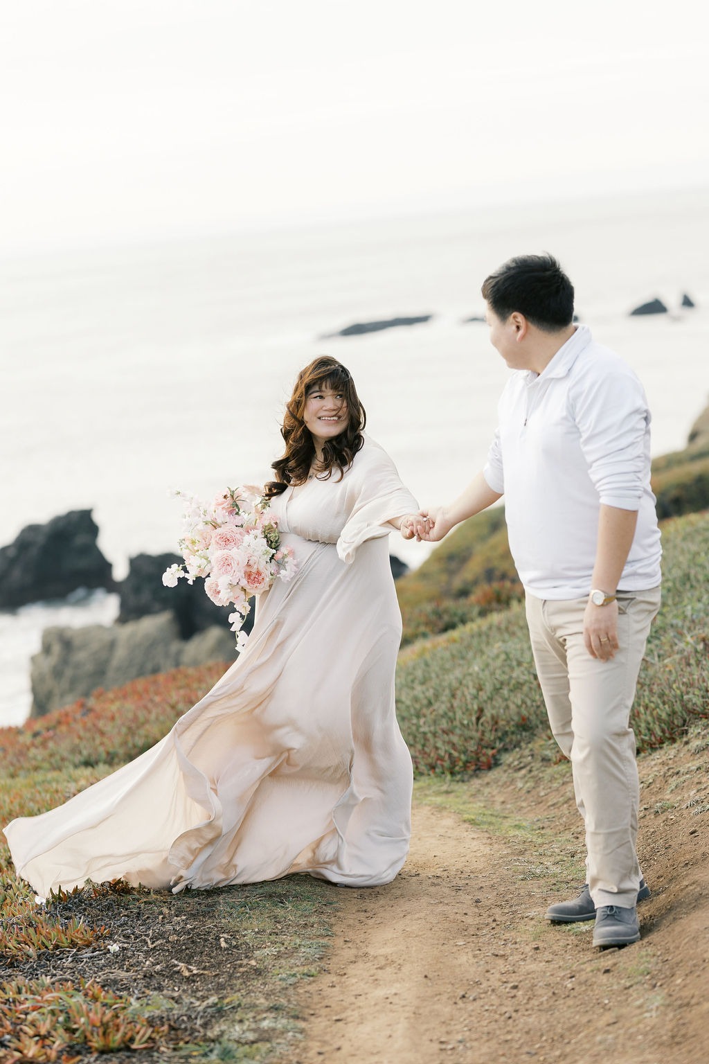 couple smiling at each other while walking on a path close to rodeo beach ca during their maternity photoshoot