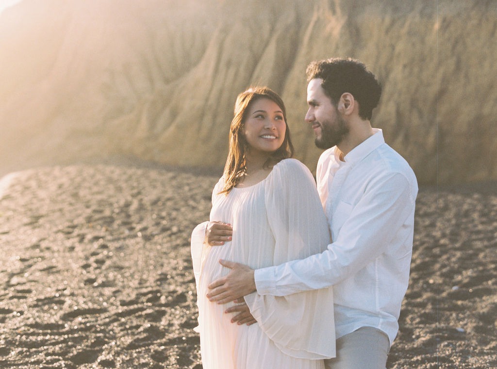 man and woman holding her pregnancy bump during soft glowy fog light while photographing at rodeo beach ca for their maternity images
