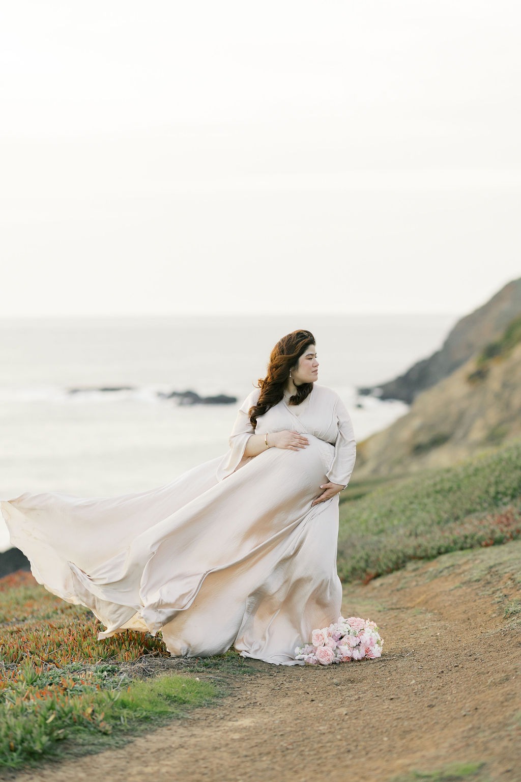 woman in a flowy champange colored dress holding her pregnancy bump during her rodeo beach san francisco ca maternity photoshoot