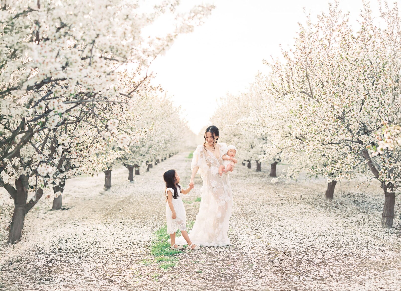 mother and daughter holding hands while the mother holds her newborn baby, standing in a field covered in white florals during their newborn family photo shoot