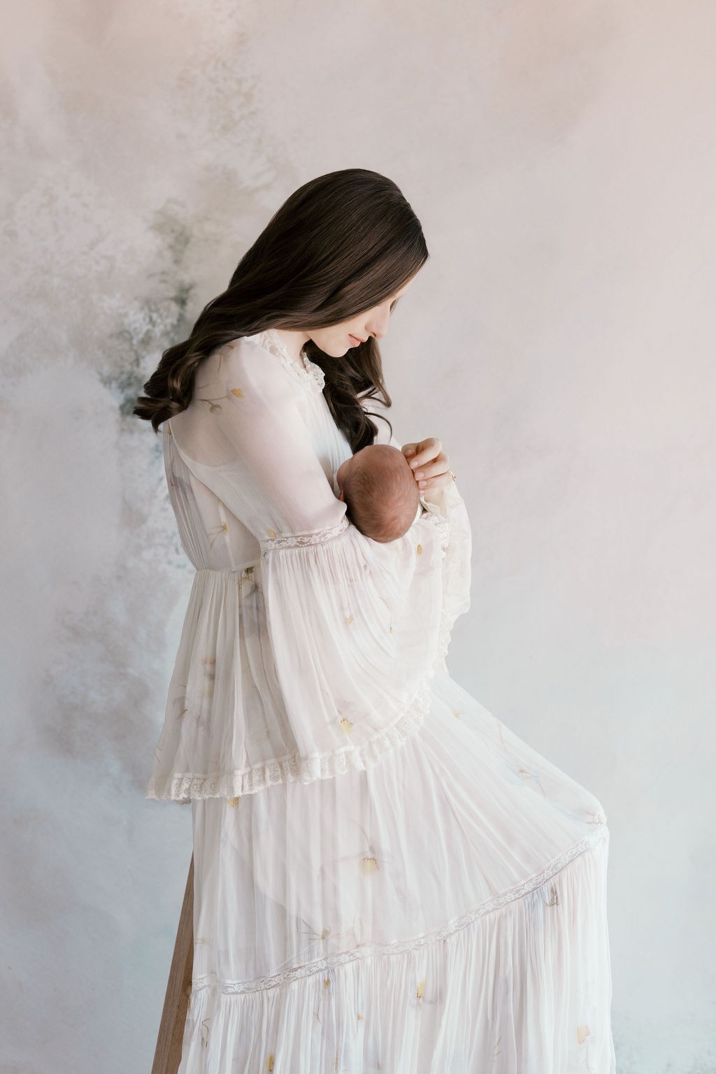 Mother in a flowing white dress looking down at her newborn while gently cradling the baby during a studio photography session