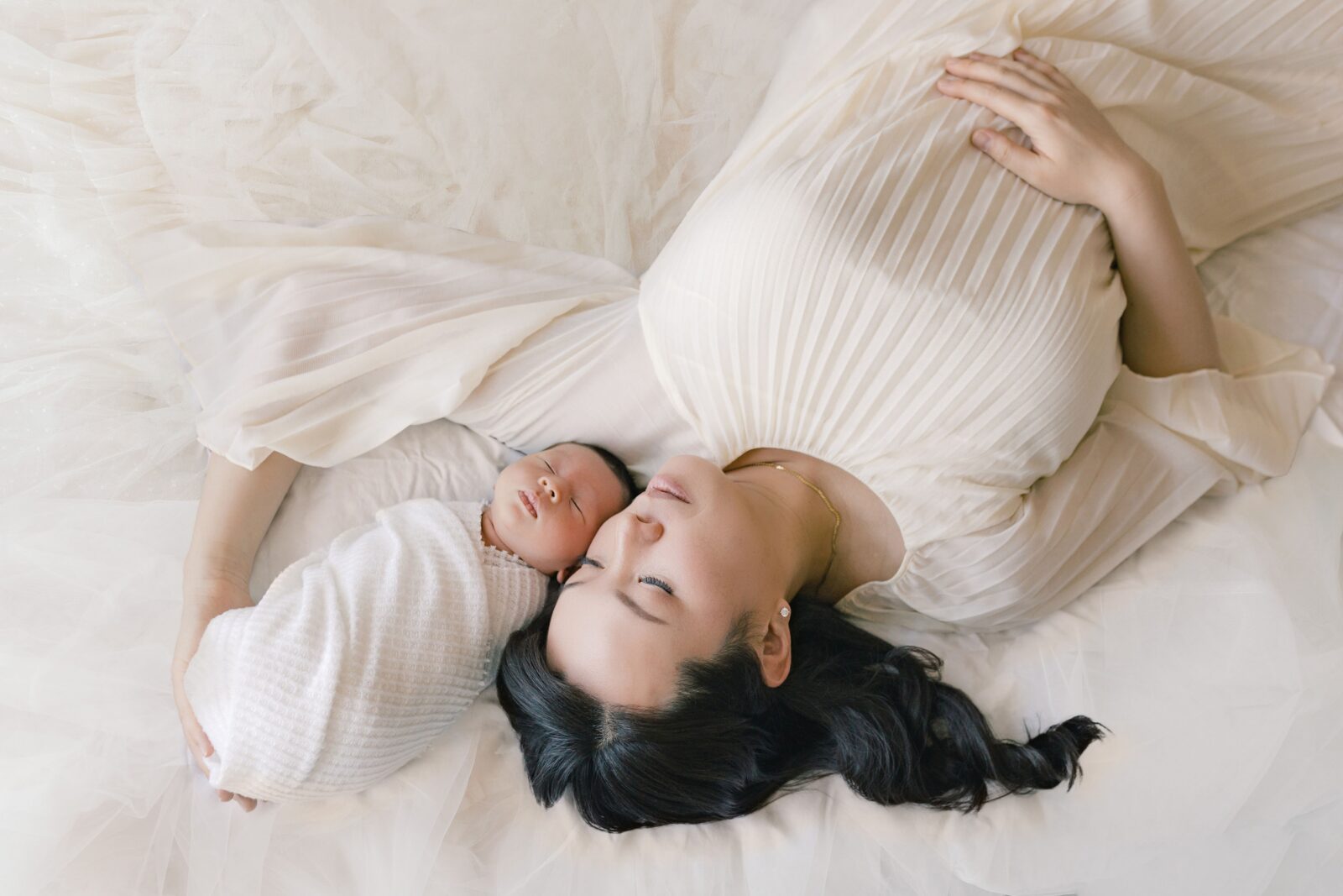 Mother lying on a white bed with her sleeping newborn nestled beside her, surrounded by soft white fabrics
