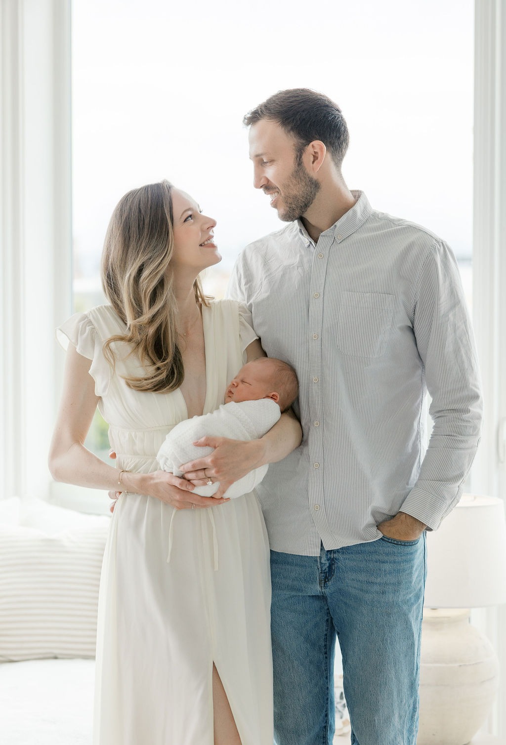 Mother holding her sleeping newborn while smiling at her partner during a bright, natural-light family photography session.